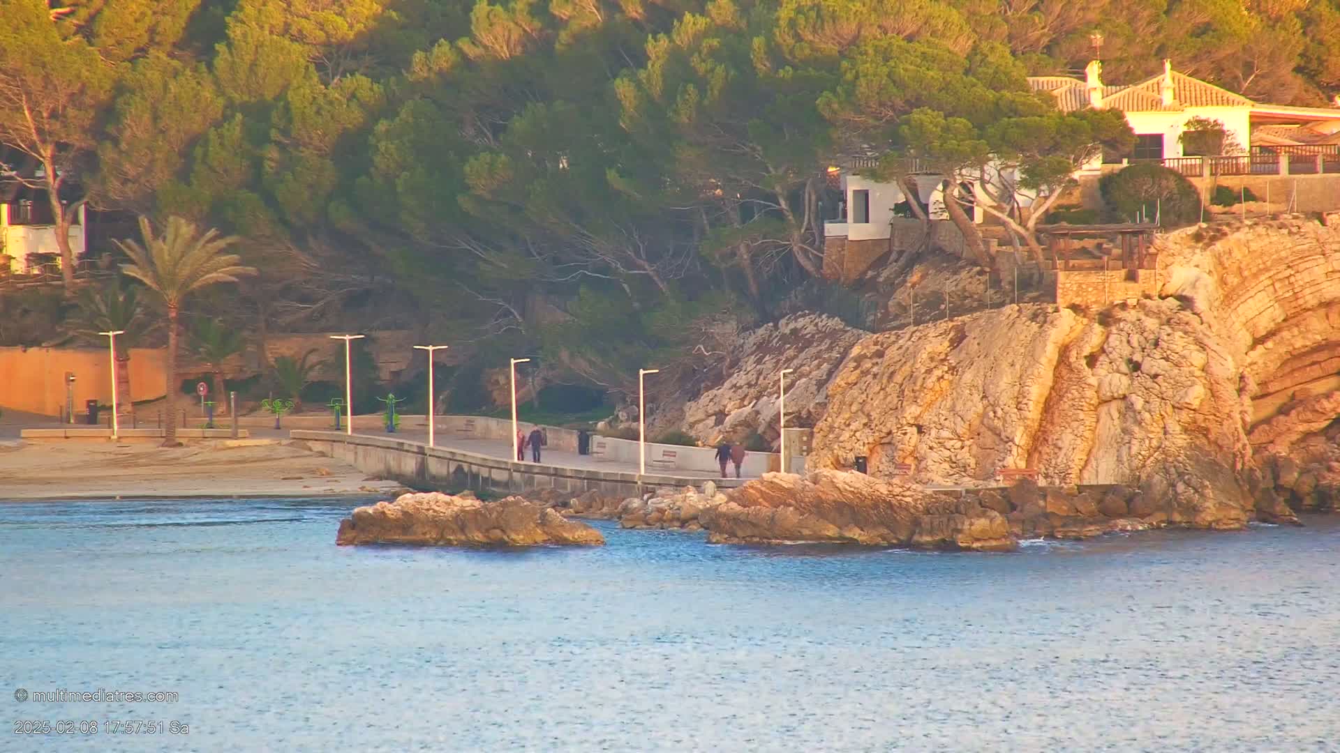 A paved walkway winds along a rocky coastline, with several people strolling along it, overlooking calm, blue water under a sunny sky.