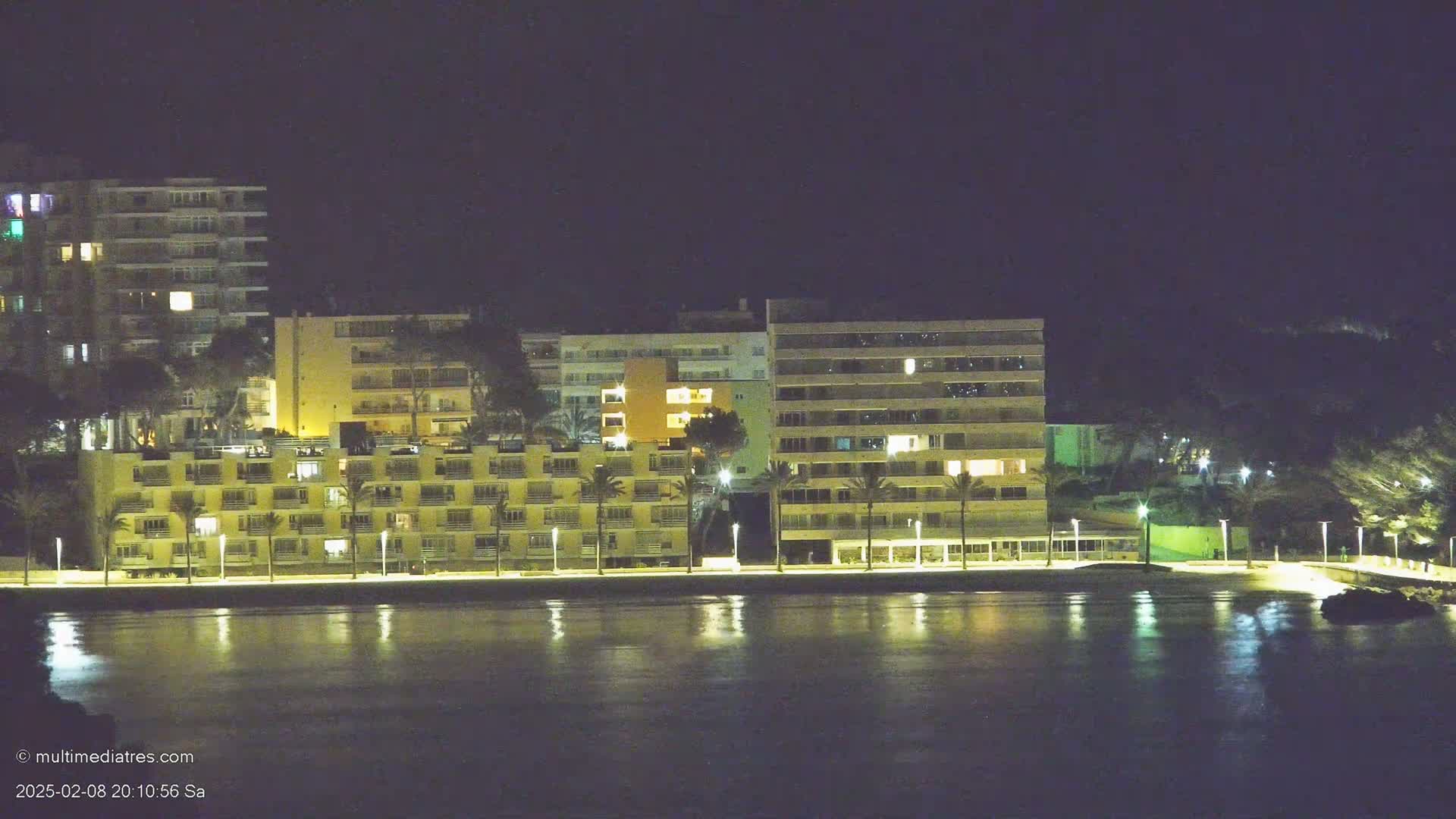 Illuminated multi-story buildings line a calm waterfront at night.