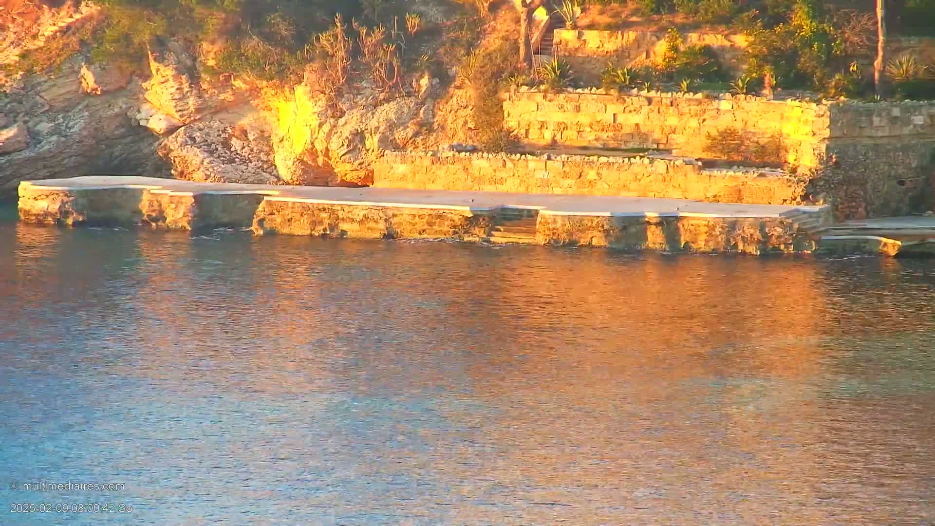 A stone platform extends into calm, golden-hued water, backed by a rocky hillside with terraced landscaping.