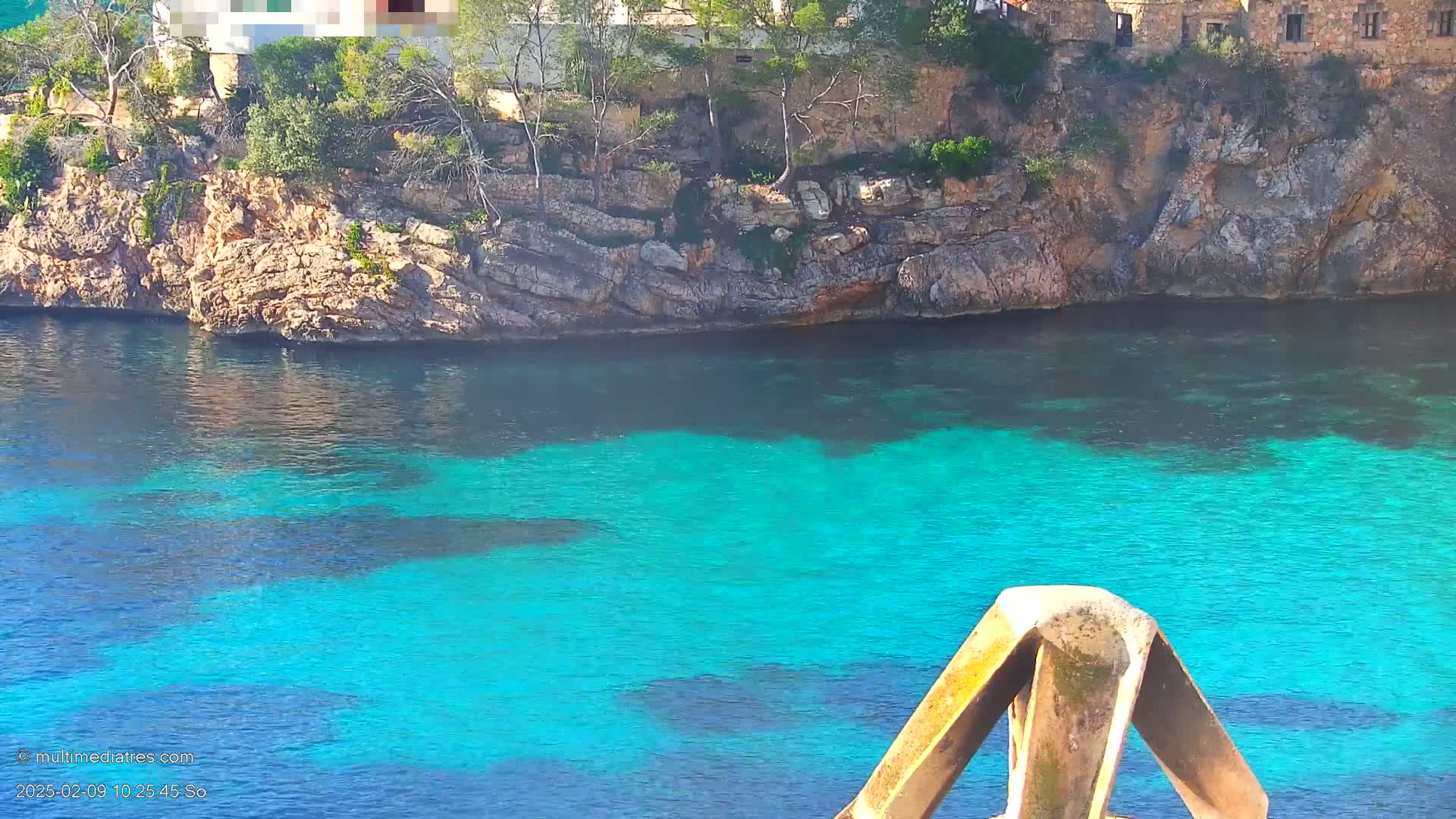 Bright turquoise water laps against a rocky shoreline dotted with trees and a stone building.