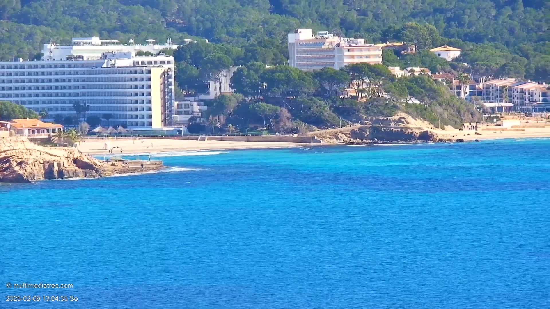 A beach with bright blue water, a sandy shoreline, and several hotels and buildings situated on a hillside covered in green trees under a sunny sky.