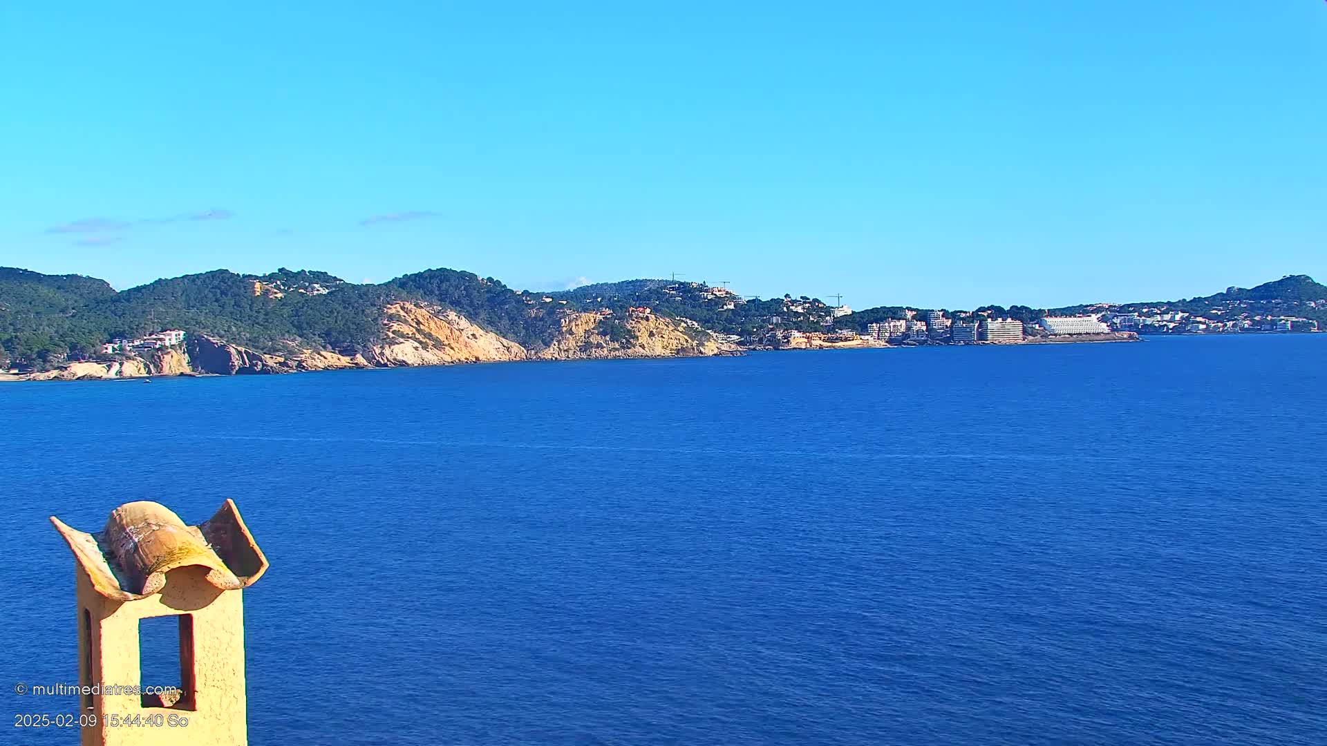 A view from atop a chimney shows a calm, blue sea under a clear blue sky, with a coastline featuring green hills, and buildings in the distance.