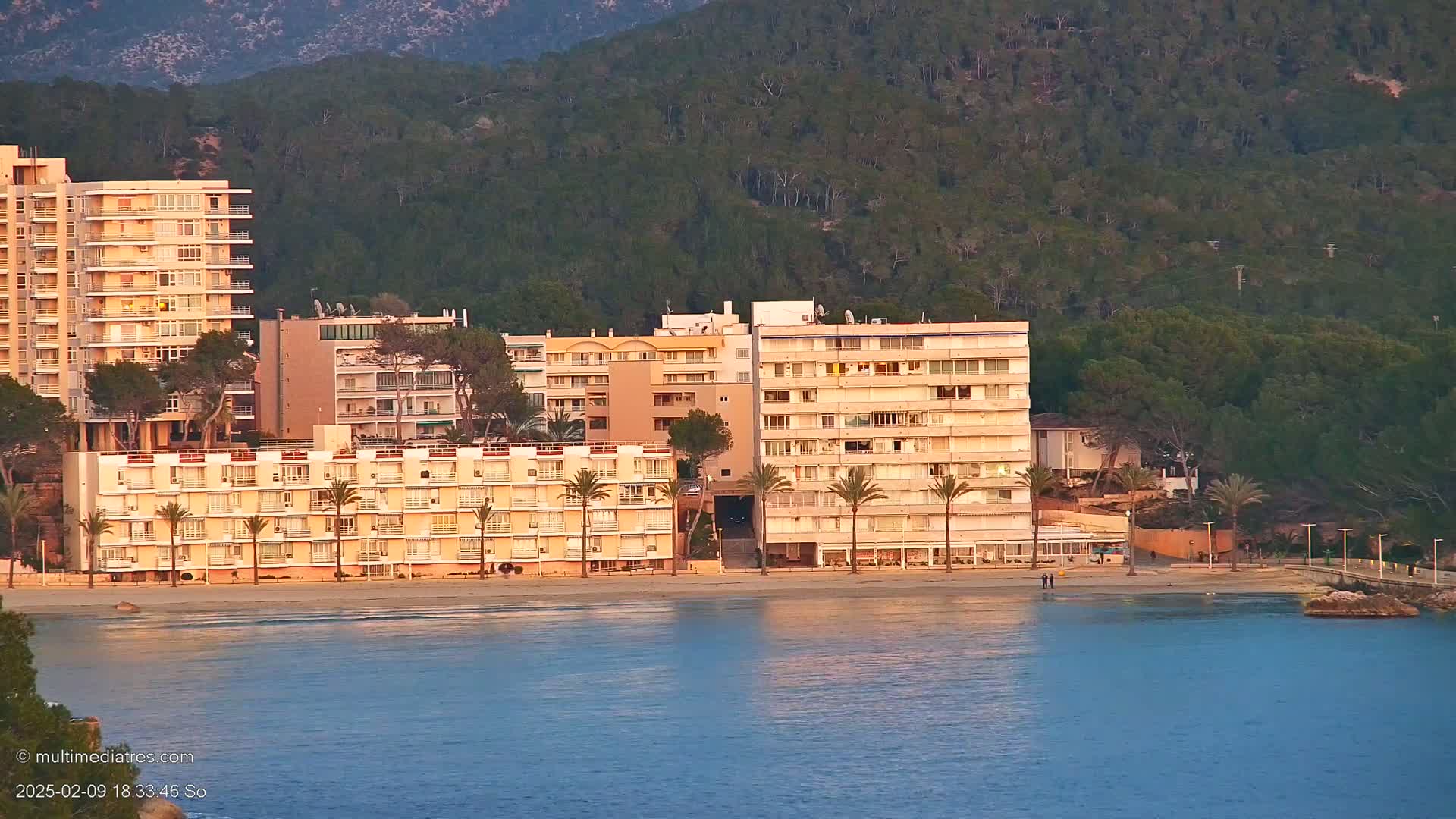 Several multi-story apartment buildings line a sandy beach next to calm water, with a hill covered in dark green trees in the background under a soft, warm-toned light.