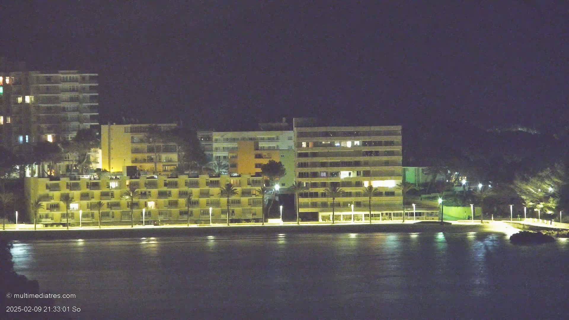 A row of illuminated apartment buildings lines a dark, calm body of water at night.