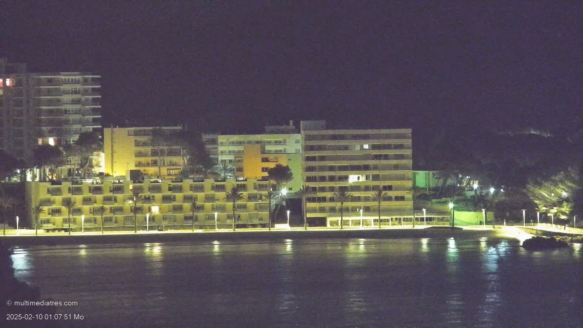 A row of illuminated apartment buildings lines a calm, dark waterfront at night.