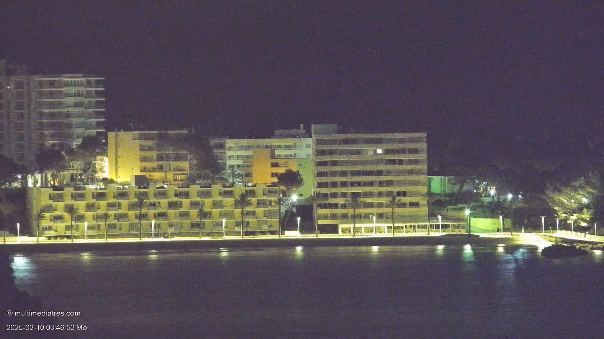 A row of illuminated beachfront buildings stands at night beside a calm body of water.