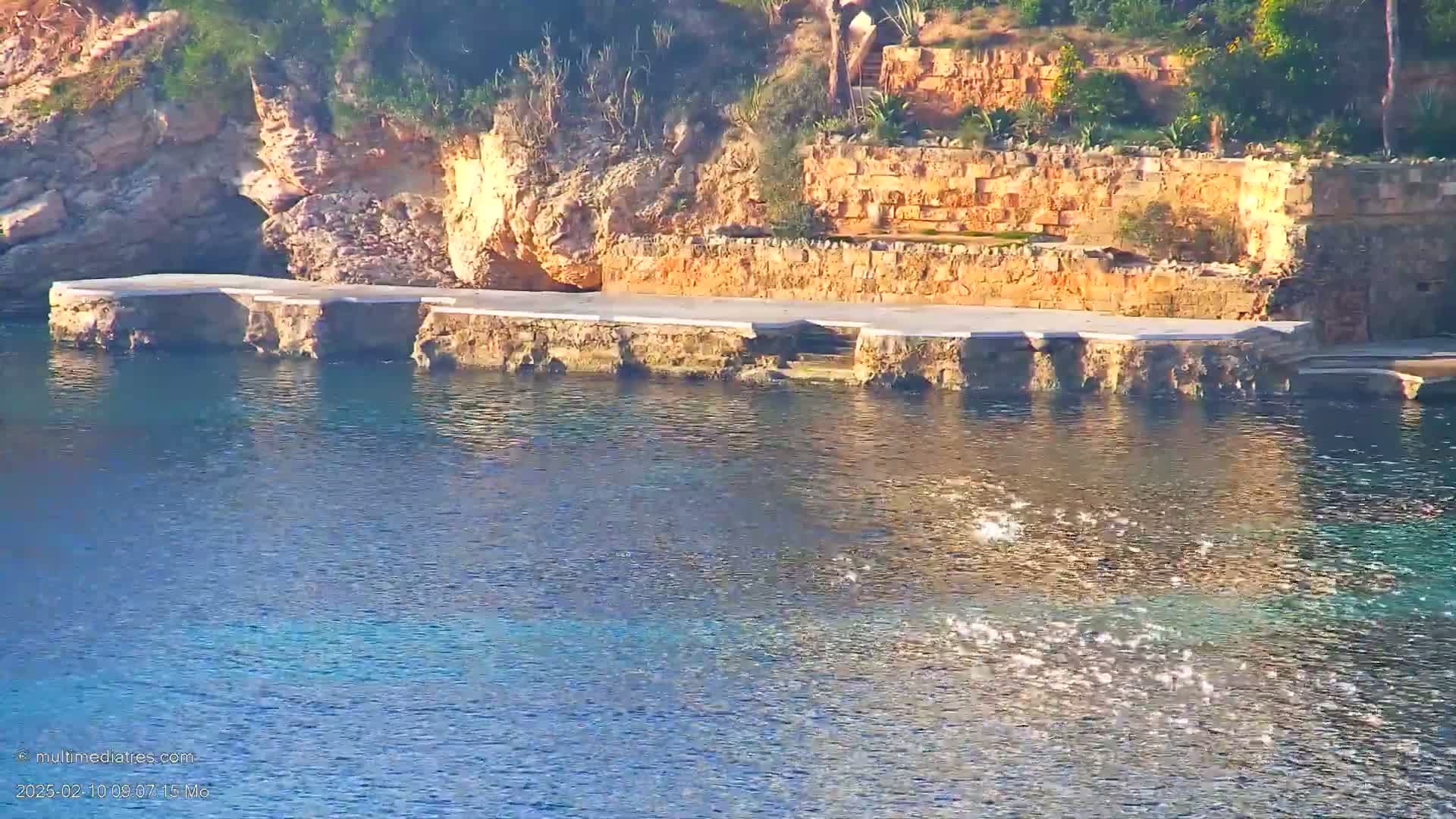 A concrete platform extends from a rocky, vegetation-covered hillside into calm, clear blue water.