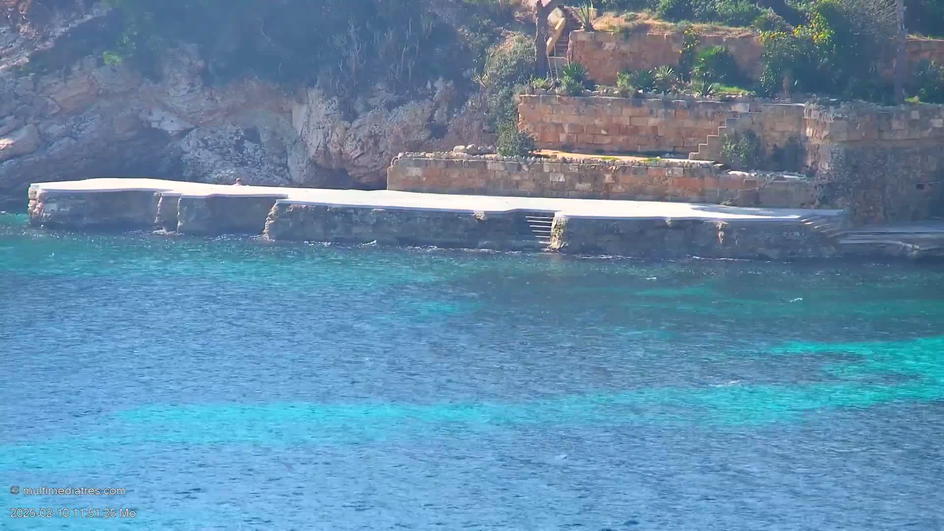 A rocky shoreline with a white concrete platform and stone steps leads down to clear, teal water under a sunny sky.
