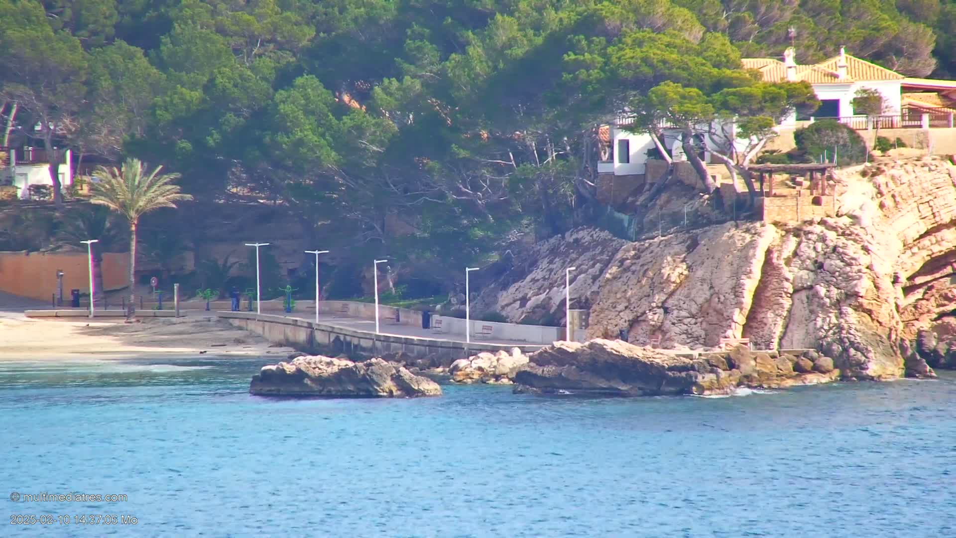 A paved walkway stretches along a rocky coastline next to a sandy beach, with several houses perched atop the cliffs under a sunny sky.
