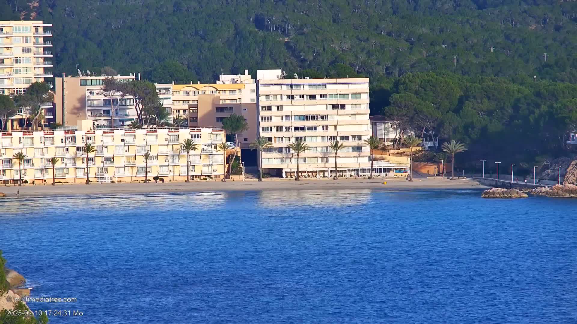 A calm blue sea stretches out in front of a sandy beach lined with multi-story buildings and palm trees, all set against a backdrop of a green hill.
