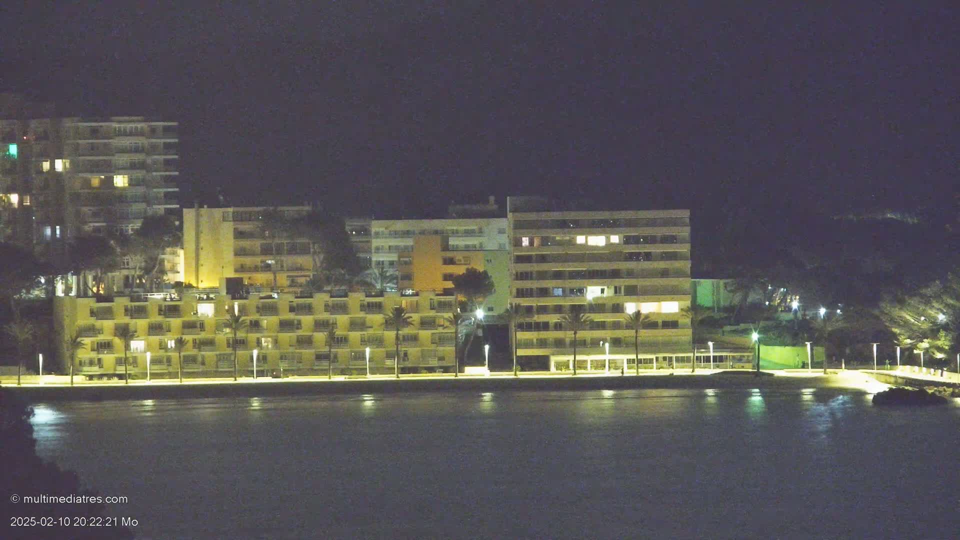 A line of illuminated beachfront buildings stands beside a dark body of water under a night sky.