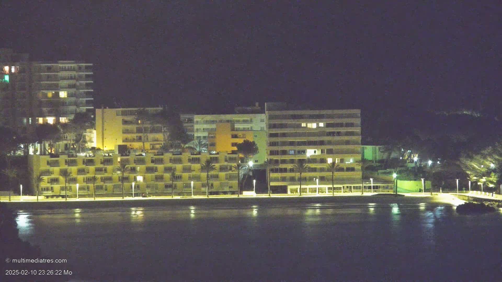 A row of illuminated multi-story buildings stands along a dark waterfront at night.