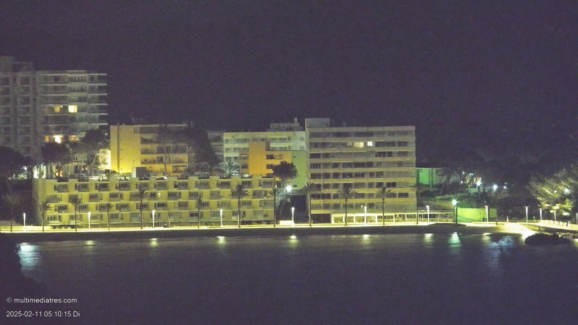 A row of lit-up apartment buildings sits along a dark waterfront at night.