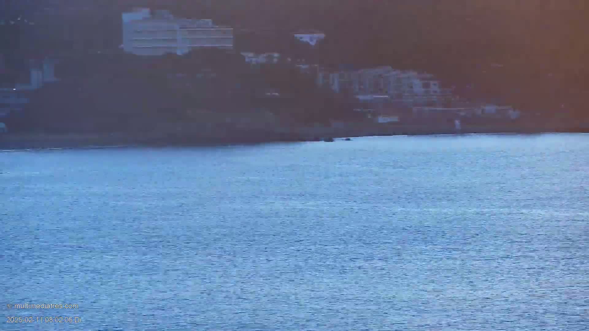 Calm blue water laps against a shoreline where several buildings are situated on a hillside.