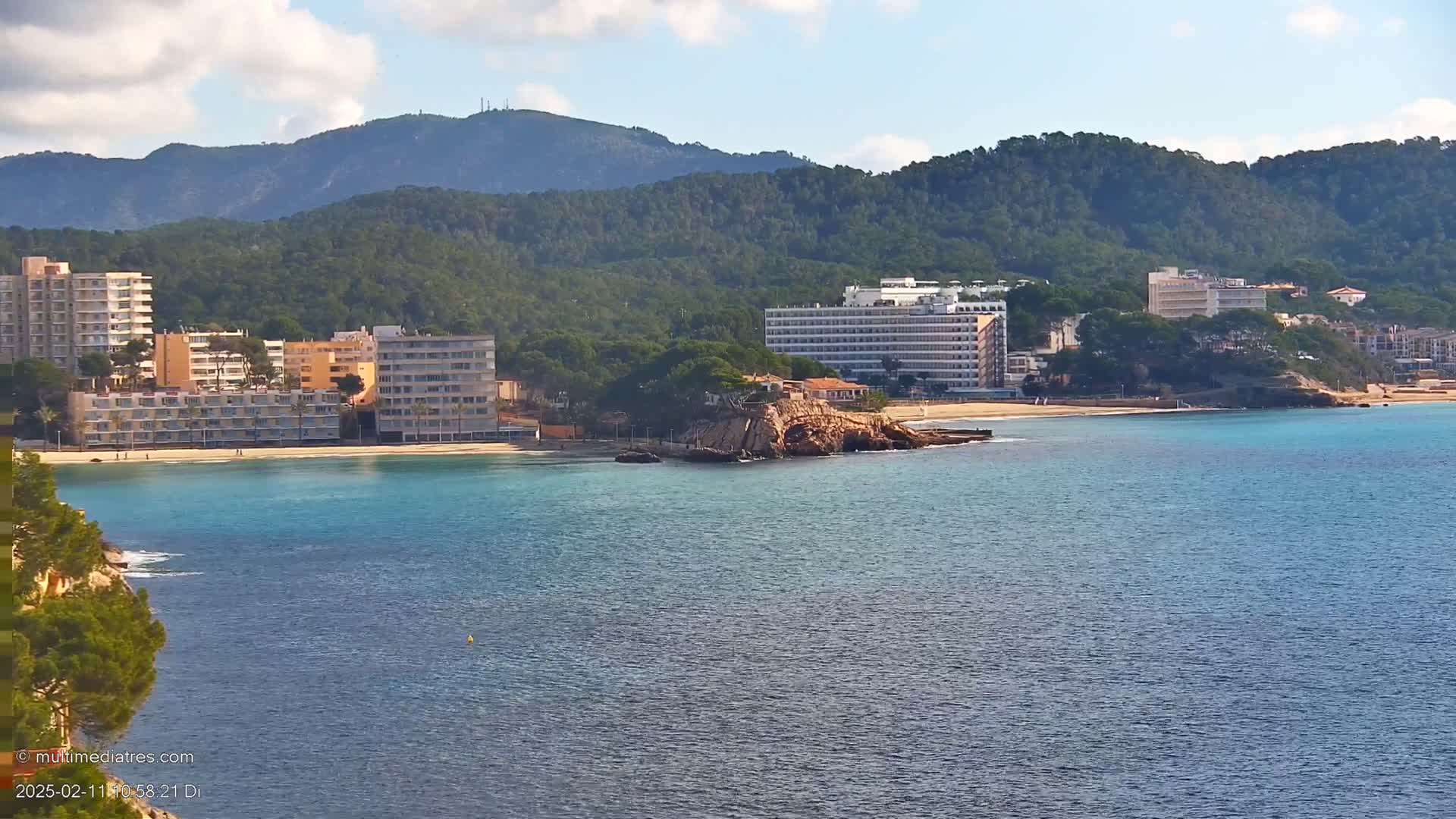 A tranquil bay with several hotels along a sandy beach, backed by lush green hills under a partly cloudy sky.