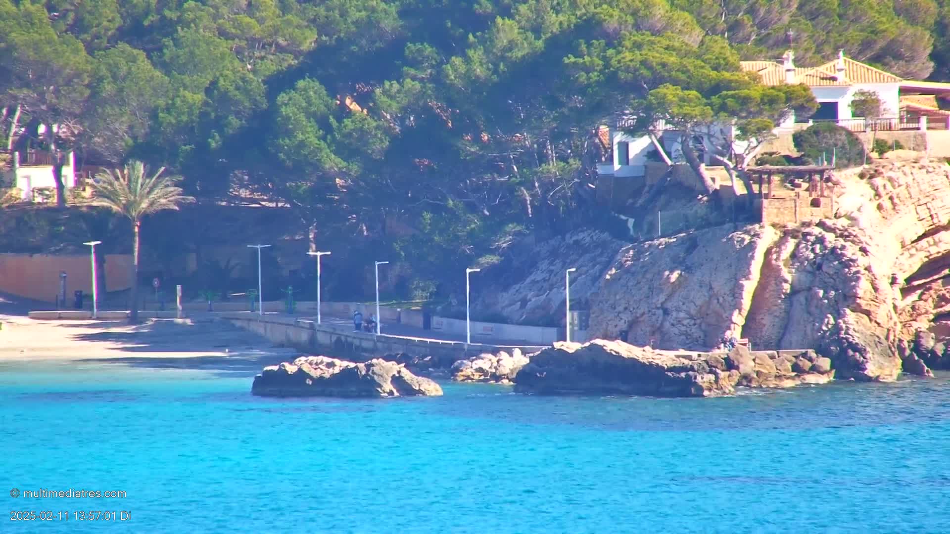 A rocky coastline with a small sandy beach, several buildings nestled amongst green vegetation, and a bright blue, calm sea.