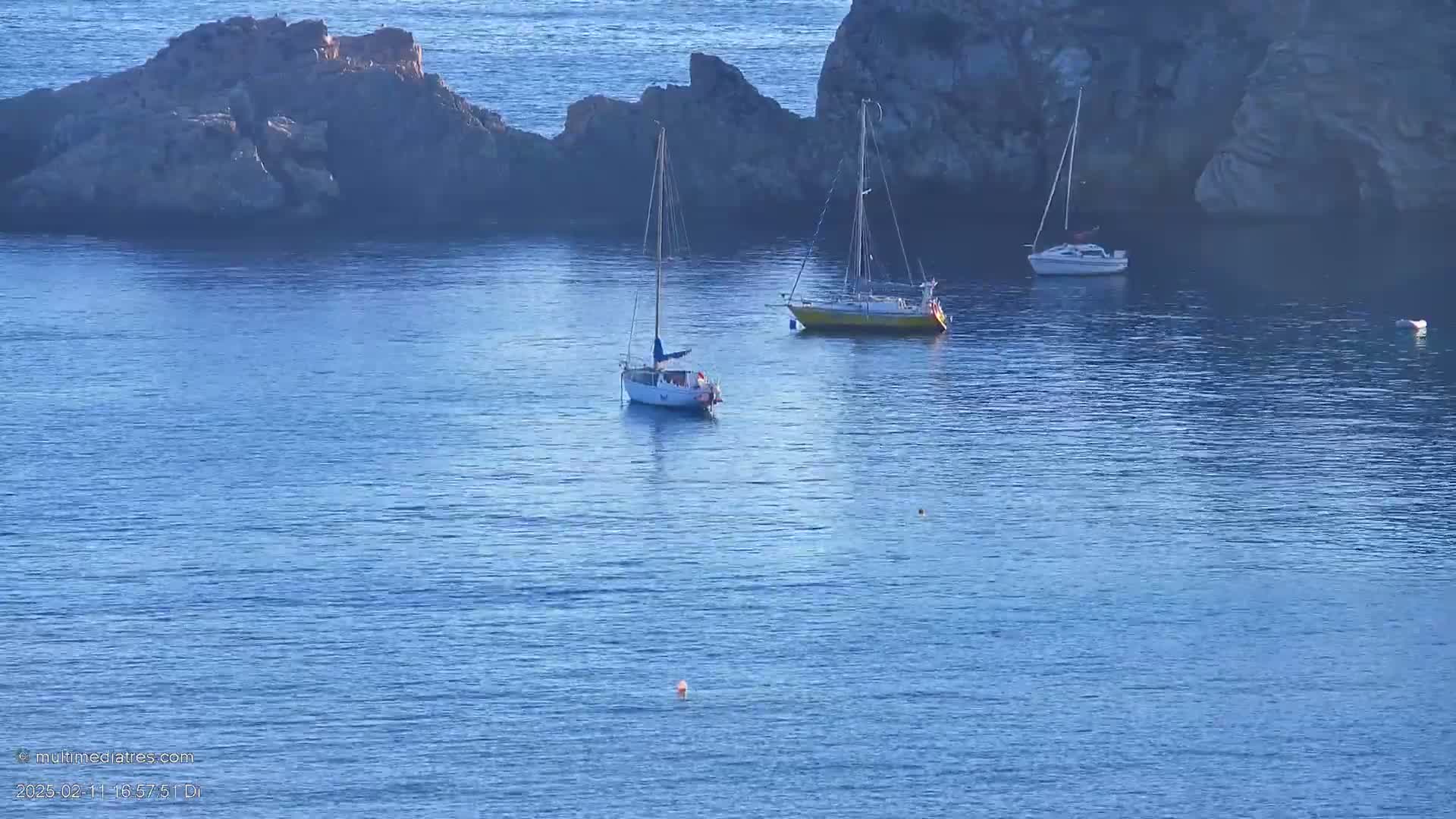 Three sailboats are anchored in calm blue water near rocky outcroppings.