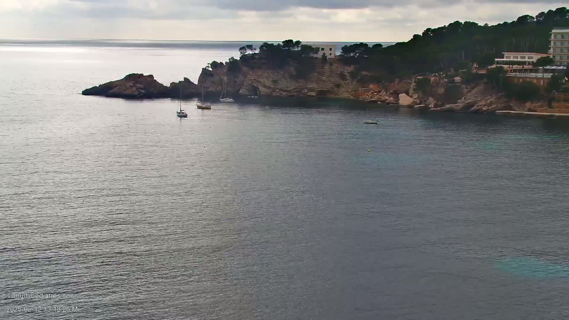 A calm, gray-sky day shows a rocky cove with a few sailboats at anchor, a building on a cliff, and dark green trees.