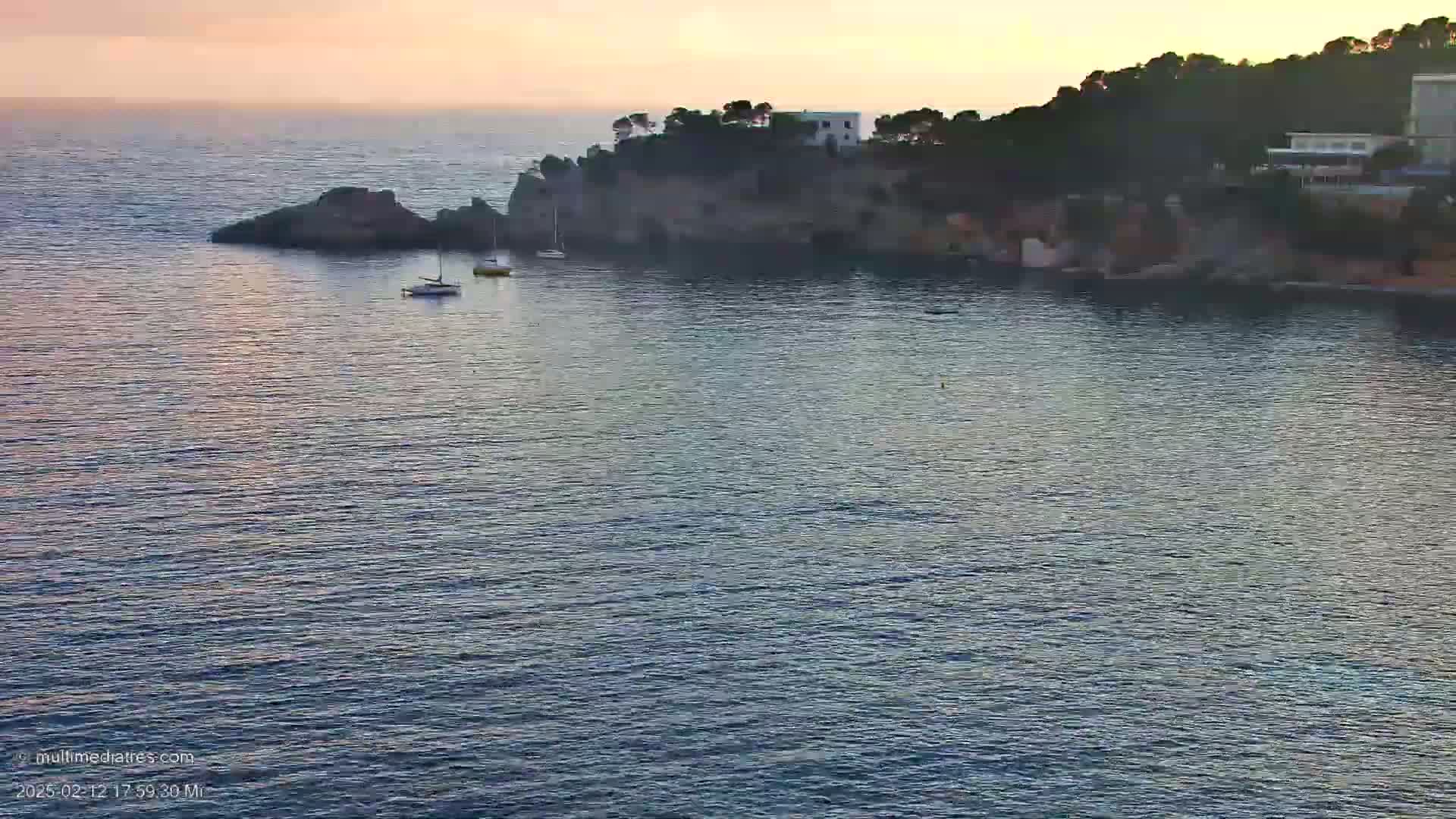 A calm, sunset-lit sea laps against a rocky shoreline where a few small boats are anchored near a cluster of buildings nestled amongst dark green trees.