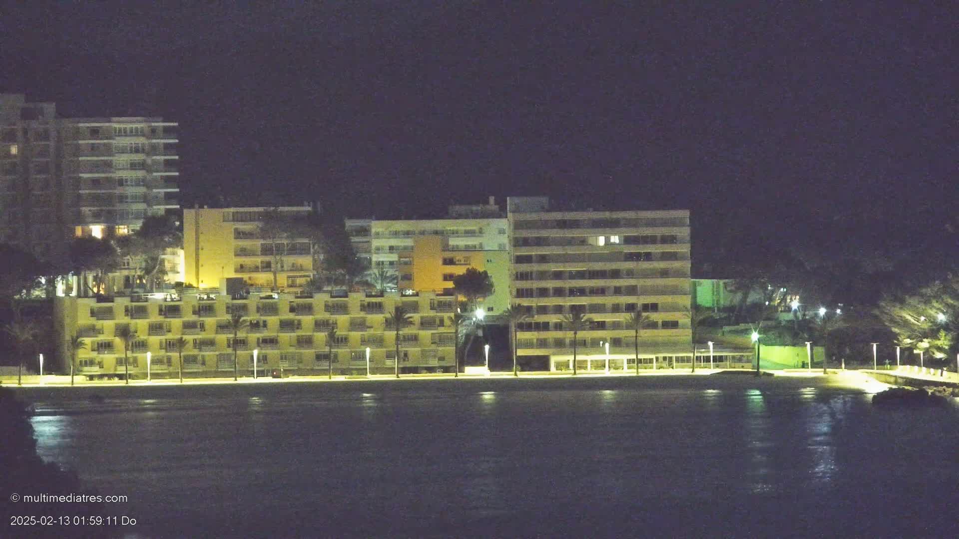 At night, several illuminated multi-story buildings line a dark beach next to a calm body of water.