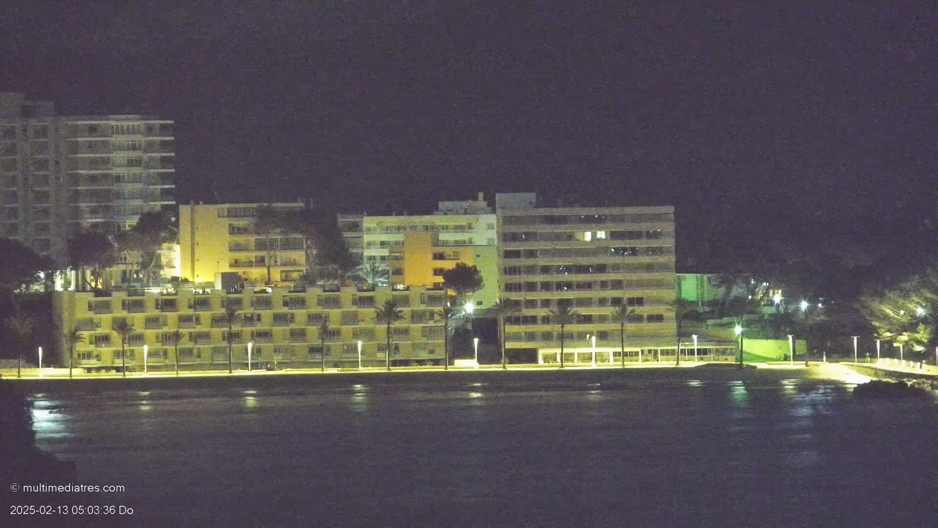 A row of illuminated apartment buildings stands along a dark waterfront at night.