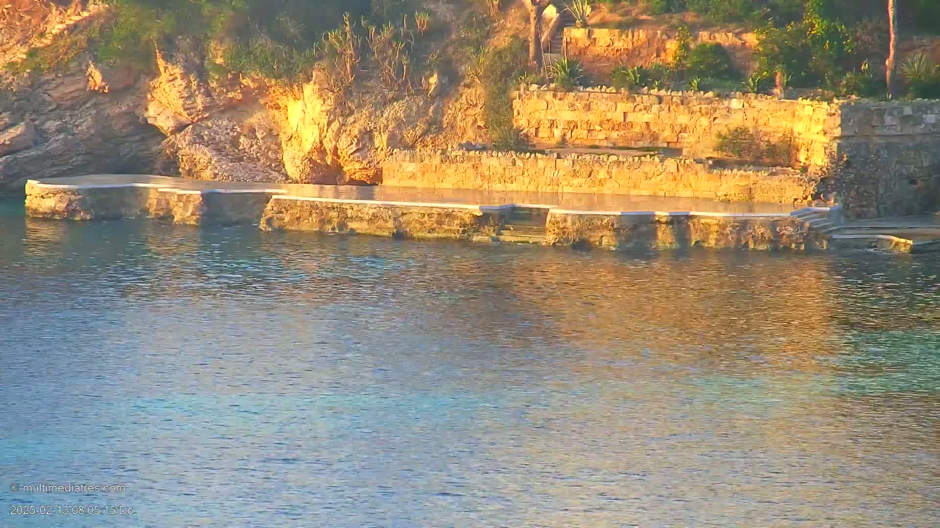 A stone platform extends from a rocky, vegetated hillside into calm, shallow, sunlit water.
