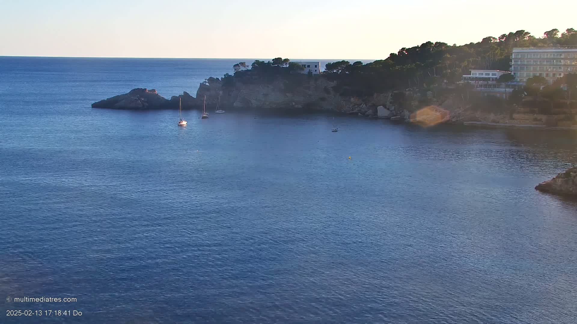 A calm, blue sea laps against a rocky coastline, where several sailboats are anchored near a small, cliffside building.