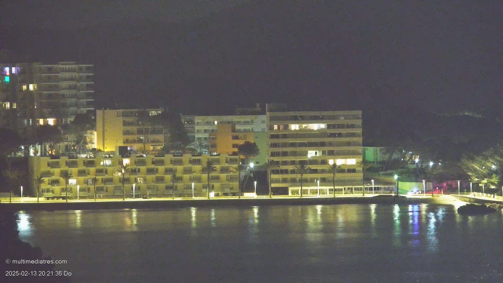 Several multi-story buildings with lights on are situated along a waterfront at night, reflecting in calm water.