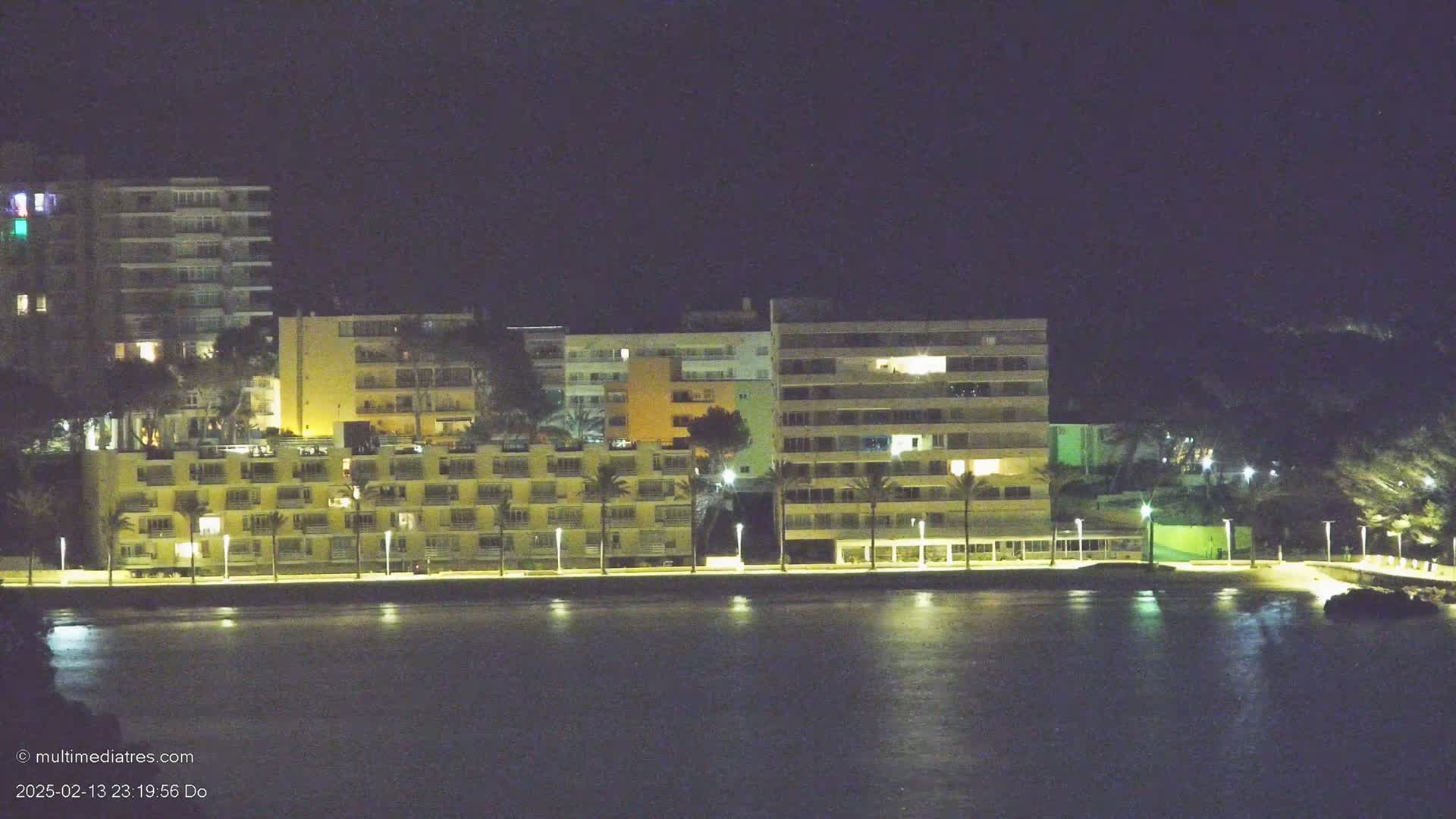 A row of illuminated apartment buildings sits along a dark, calm body of water at night.