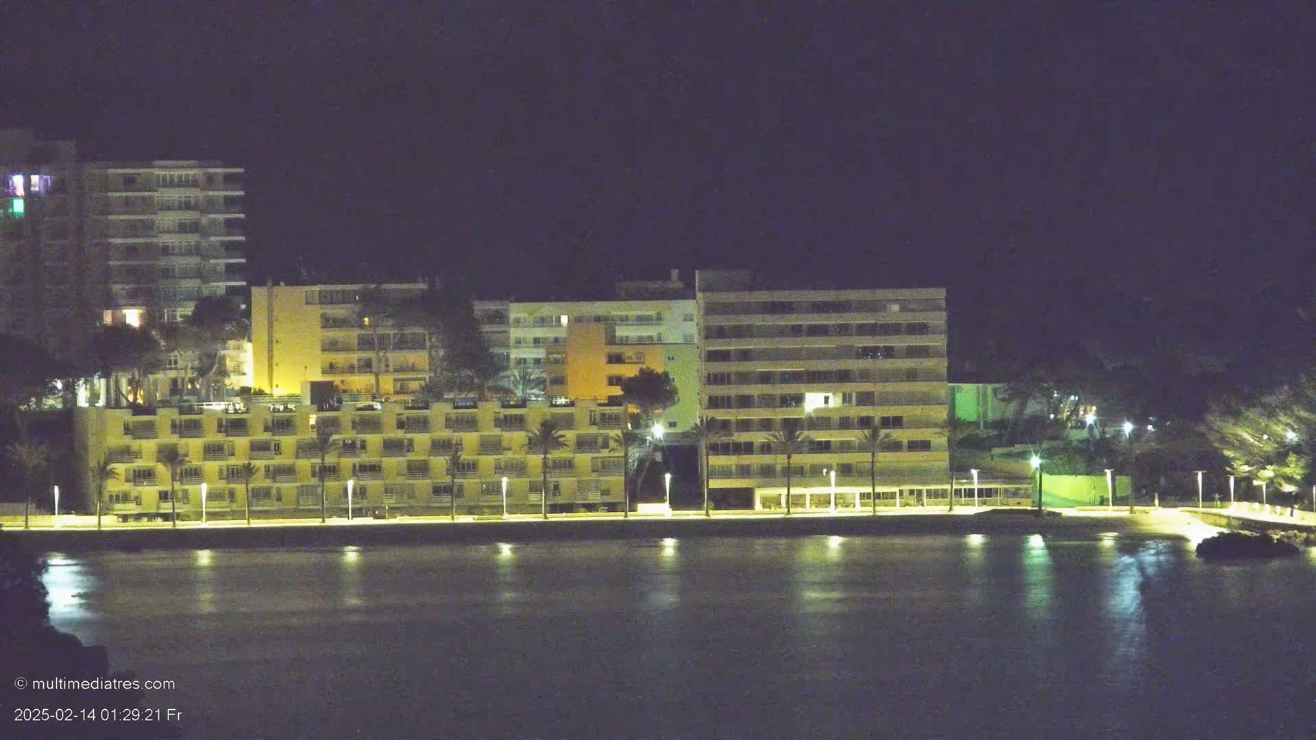 A row of illuminated beachfront hotels and buildings at night, reflected in calm water.