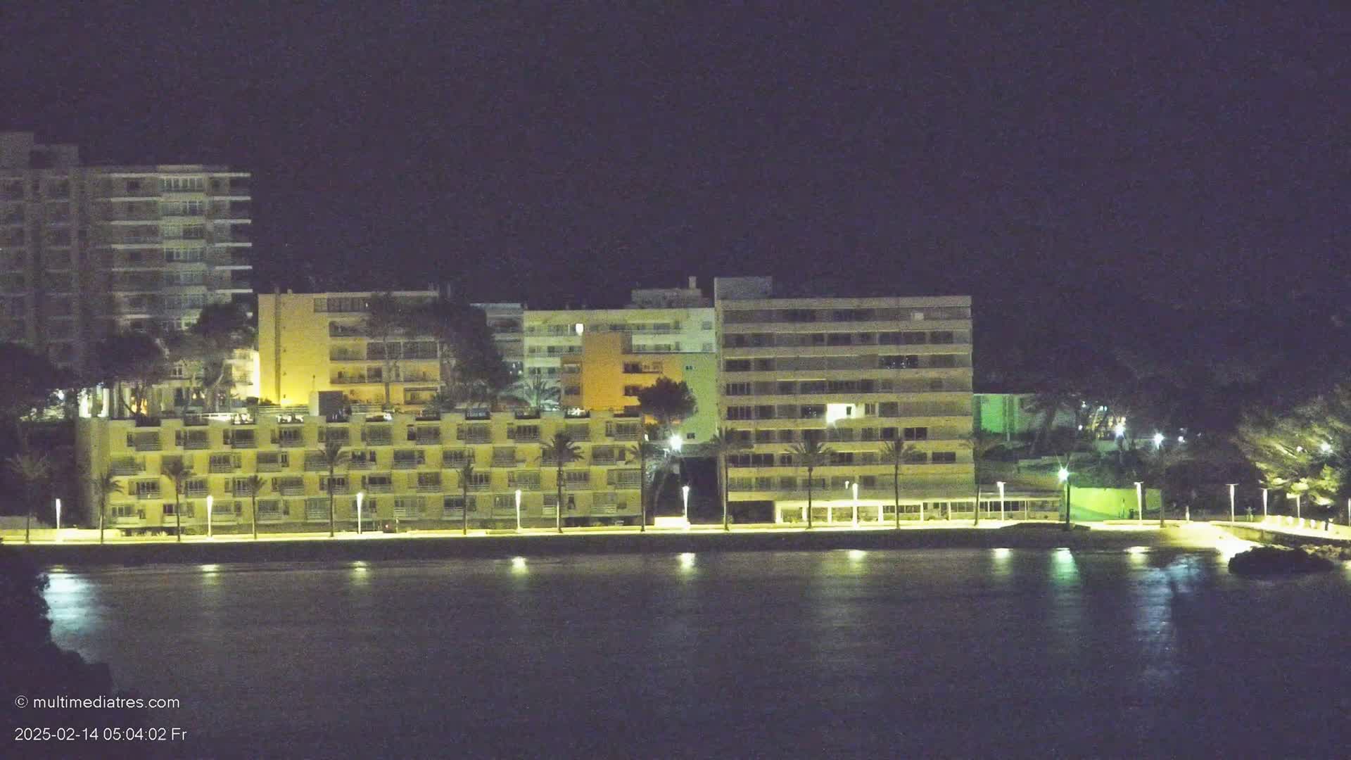 A row of lit-up buildings sits along a dark, calm body of water at night.