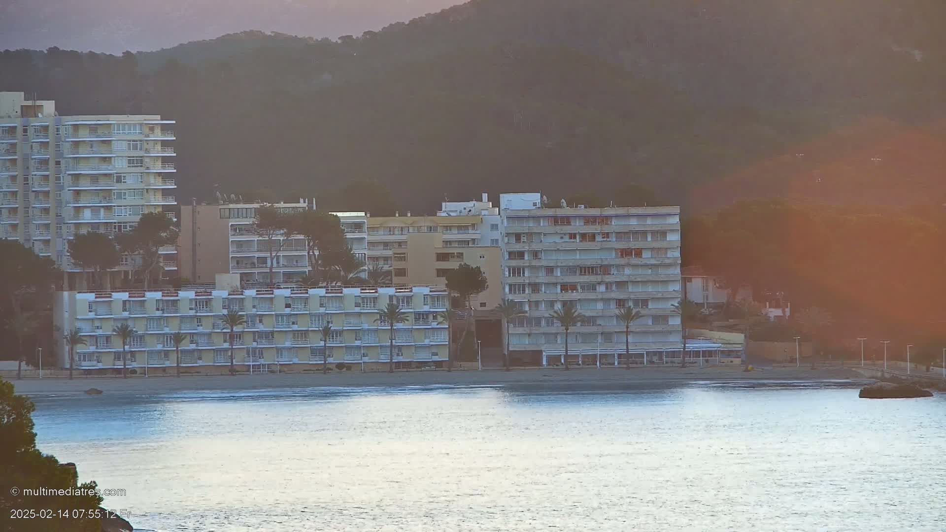 Several multi-story buildings are situated on a sandy beach next to calm water, with hills in the background under a hazy, sunrise sky.