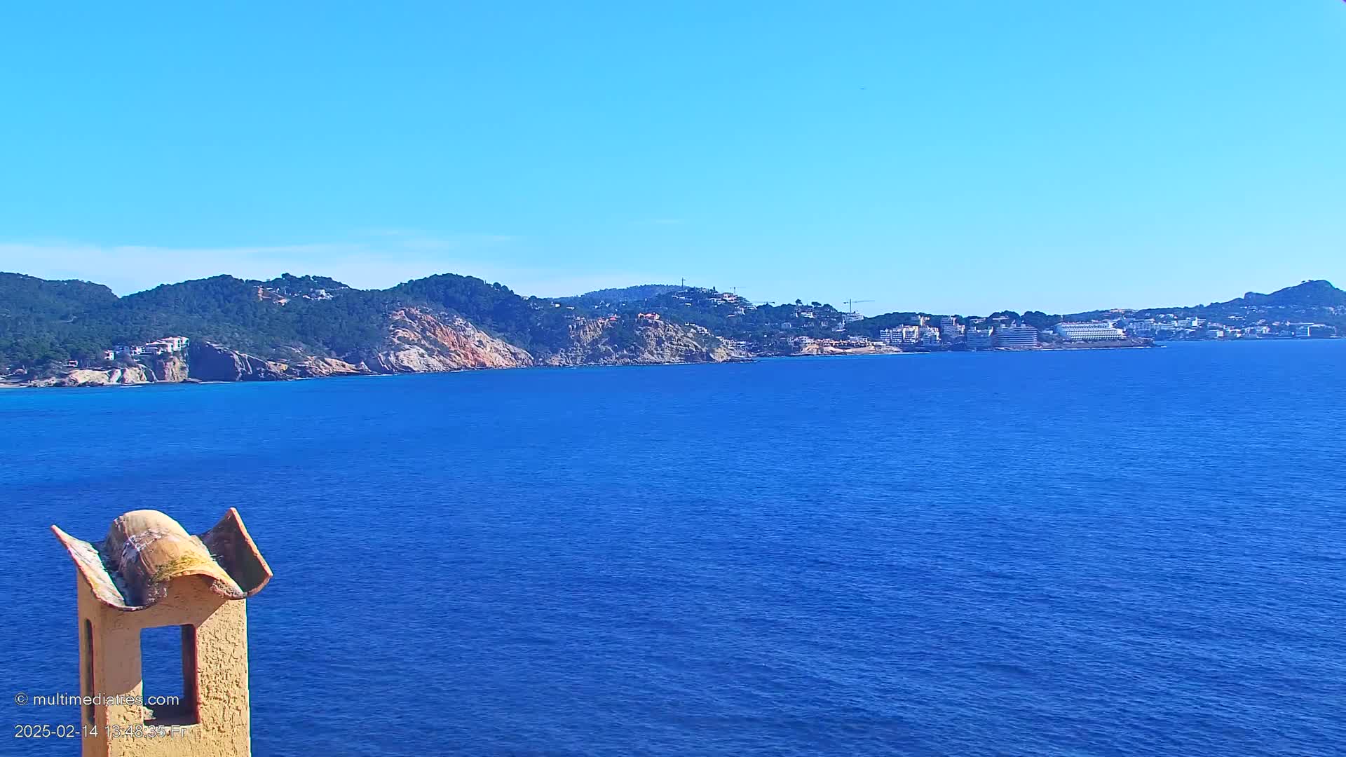 A seaside view shows a calm, deep blue ocean stretching to a distant shoreline of green hills and buildings under a clear blue sky.