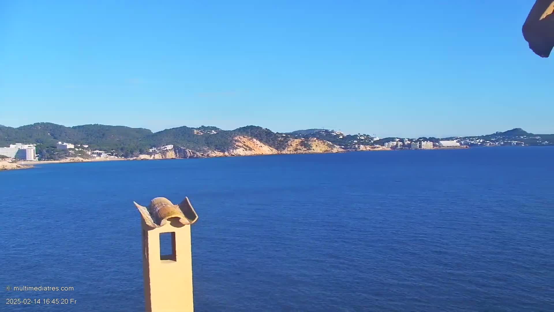 A calm, blue sea stretches before a coastline of green hills and low-rise buildings under a clear blue sky.