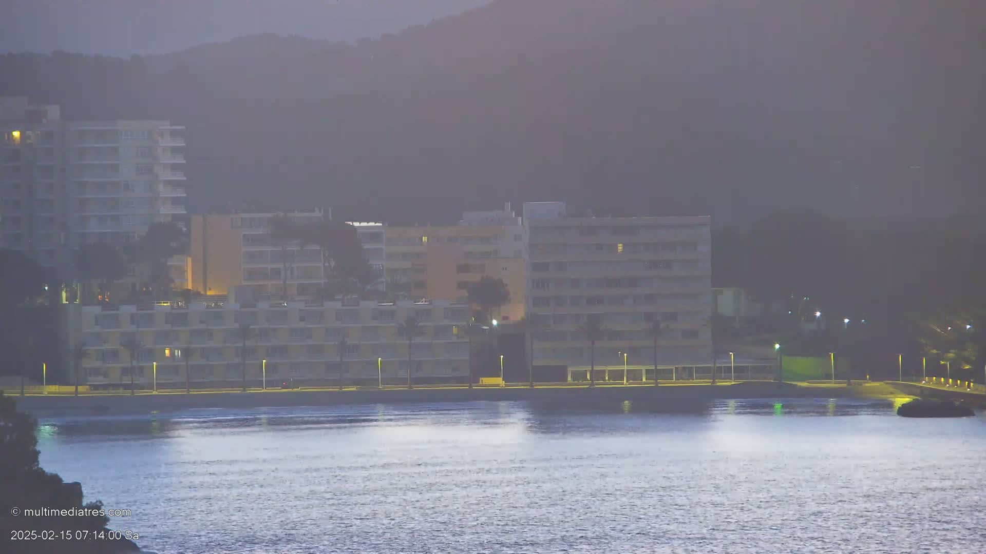 A hazy twilight scene shows several multi-story buildings along a waterfront, illuminated by lights, with calm water in the foreground.