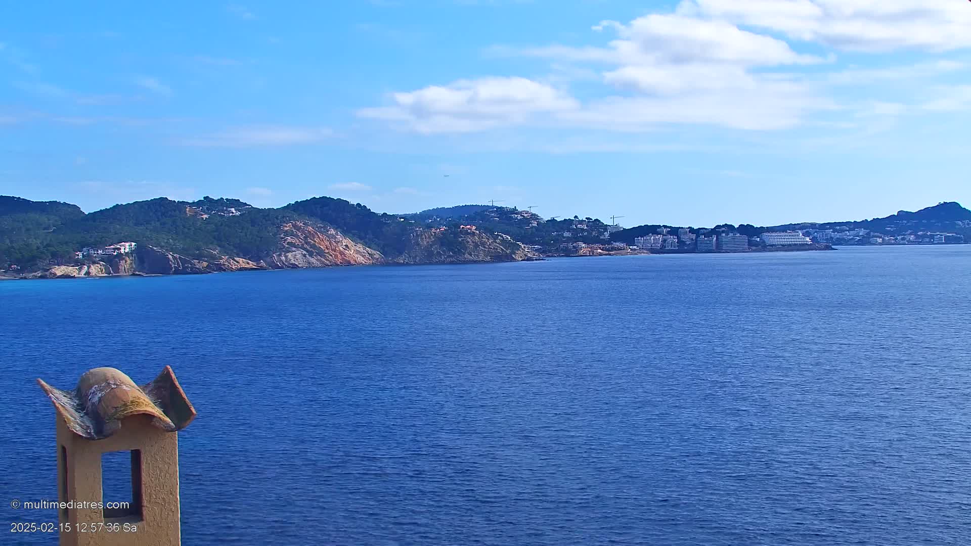 A view from atop a structure shows a calm blue sea extending to a distant coastline of rocky hills and buildings under a mostly clear sky.