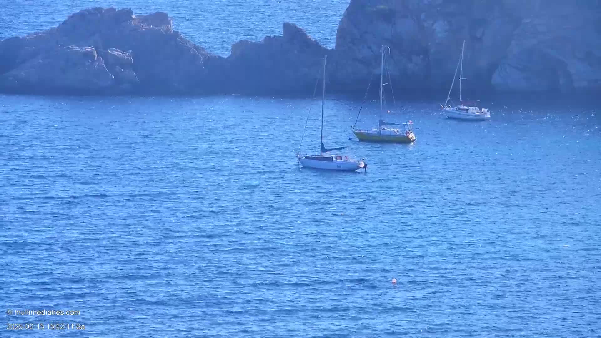 Three sailboats are anchored in calm blue water near a rocky shoreline.