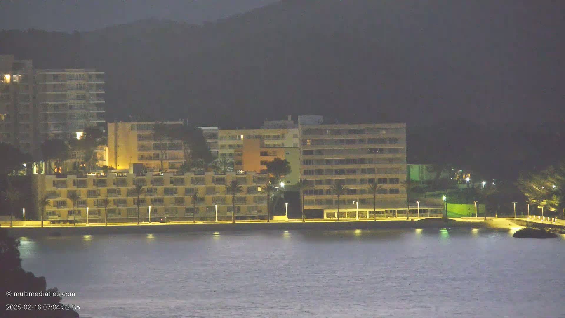 A row of lit apartment buildings sits along a dark, calm body of water at night, with a hill rising behind them.