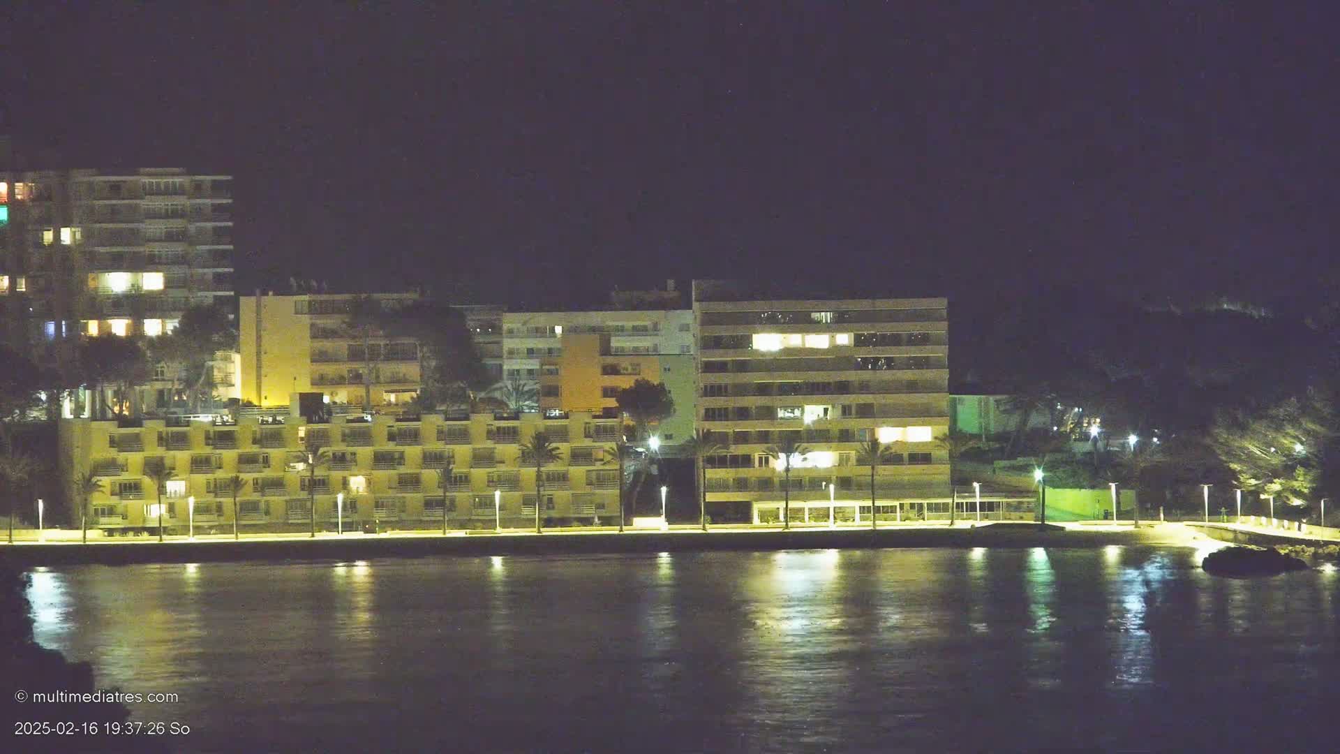 Several illuminated buildings line a waterfront at night, their lights reflecting on calm water.