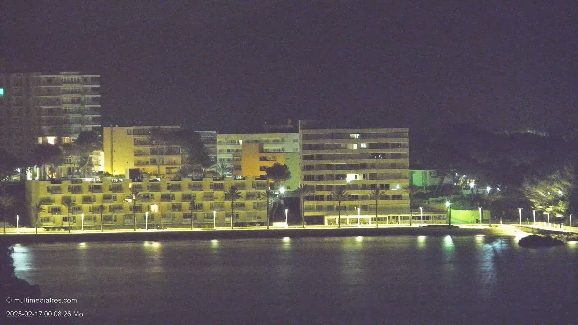 A line of illuminated multi-story buildings sits along a dark waterfront at night.
