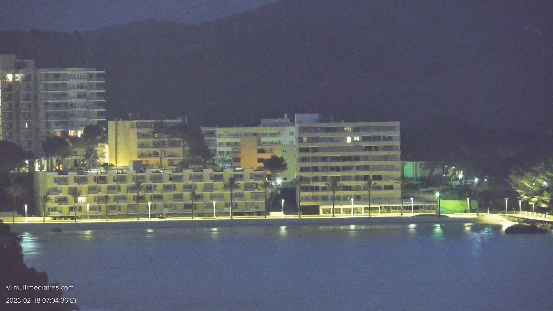 A row of illuminated beachfront buildings sits beside a dark body of water under a night sky.