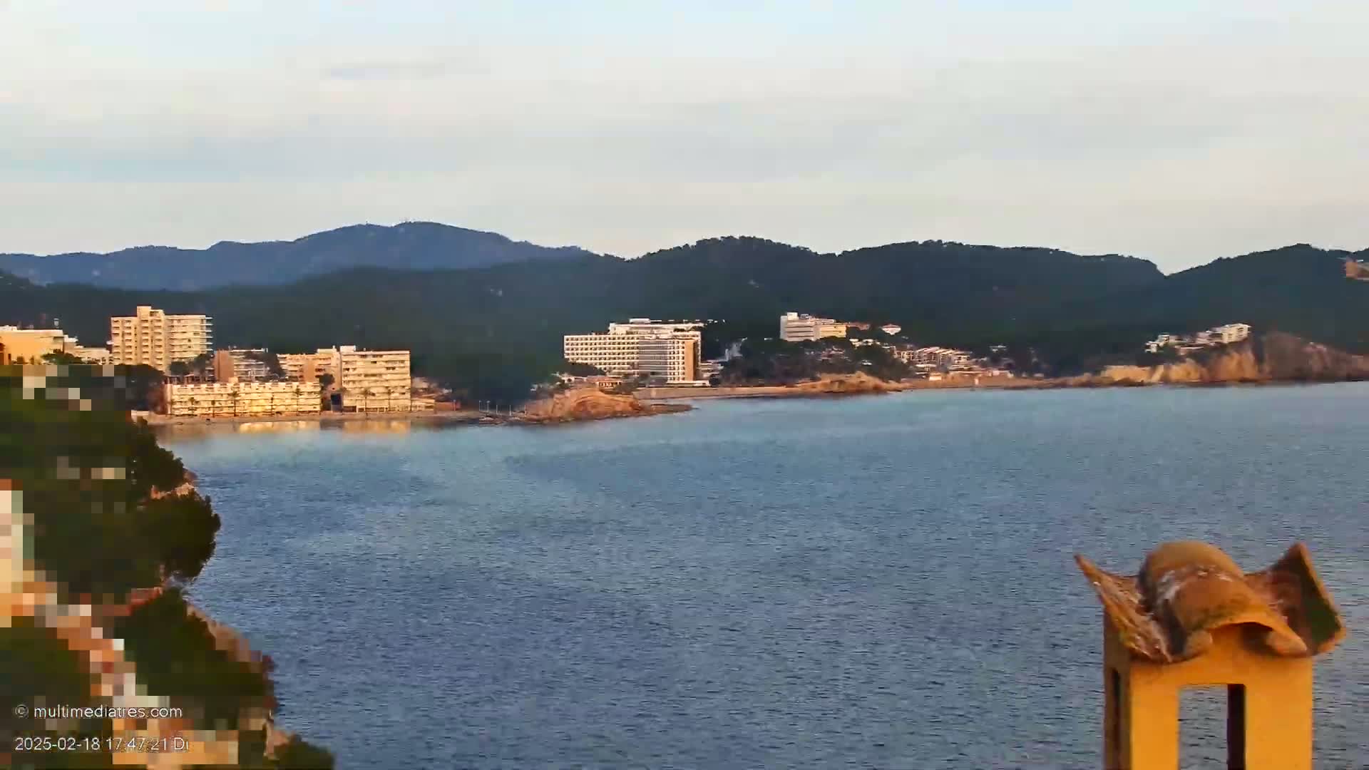 A coastal scene shows calm water separating a rocky shoreline with hotels and buildings from dark green hills under a mostly clear sky.