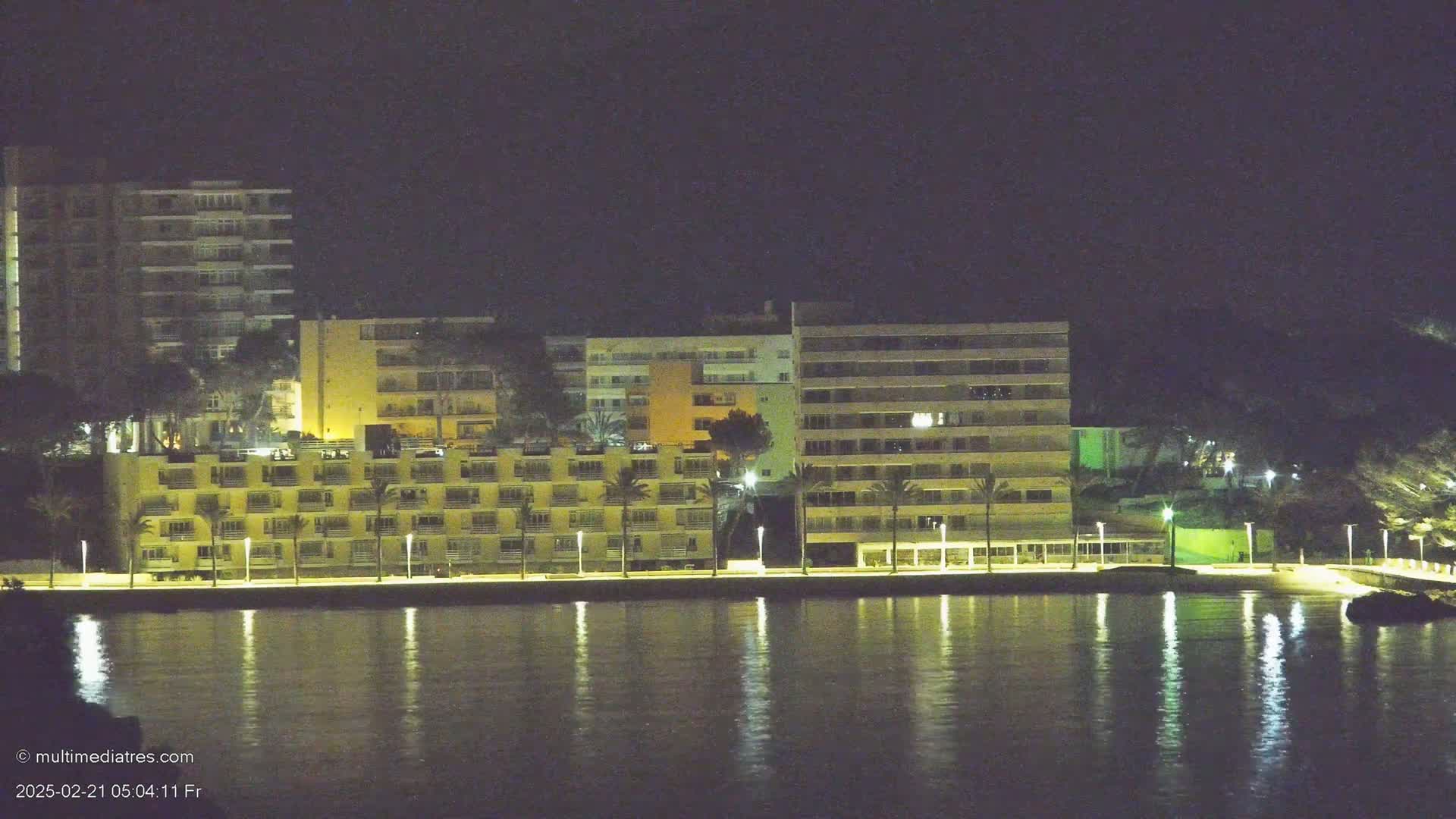 Several multi-story buildings with lights on are situated along a waterfront at night, their reflections shimmering in the calm water.