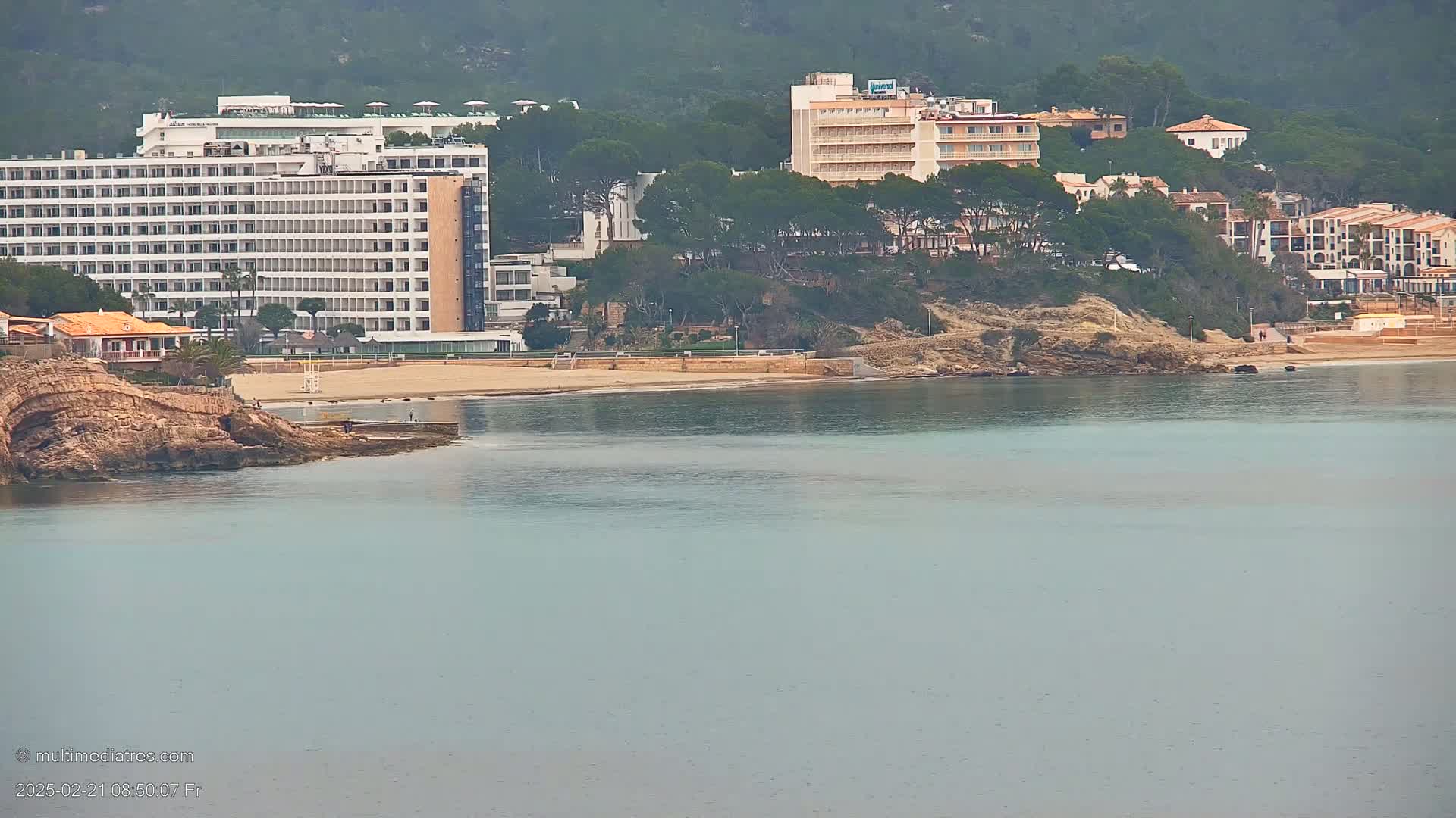 A calm, light-blue sea laps a sandy beach lined with hotels and buildings nestled amongst green hills under an overcast sky.