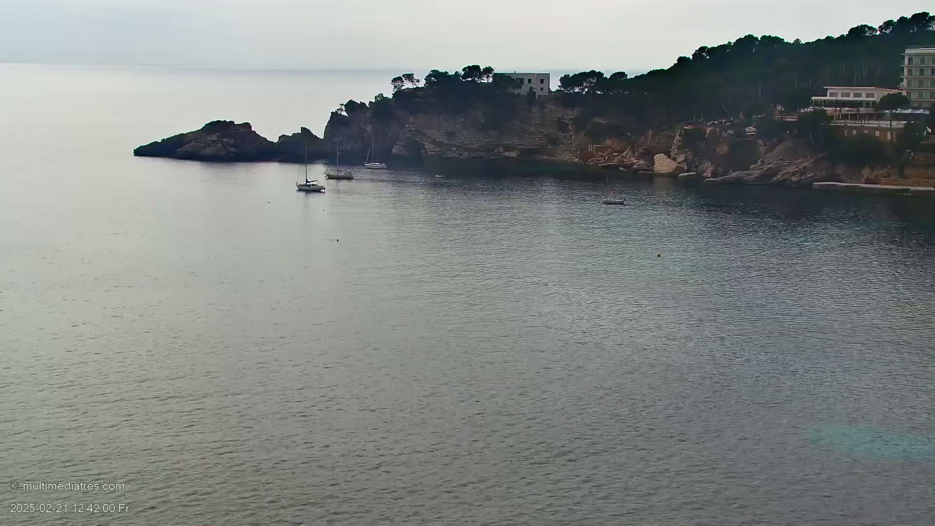 A calm, gray-toned sea laps against a rocky shoreline where a few sailboats are anchored near a small, tree-covered peninsula with a building on top.