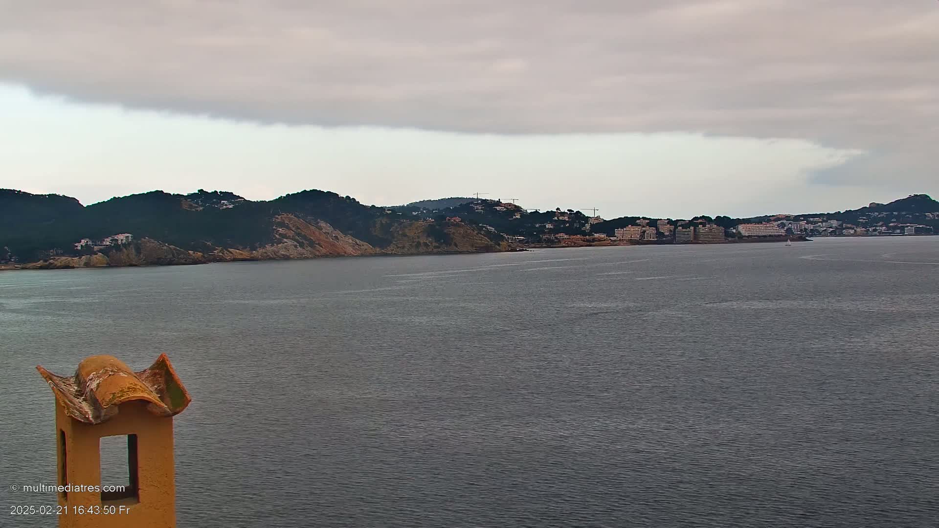 A view from a rooftop shows a calm, gray body of water stretching to a distant coastline of dark green hills and low-rise buildings under an overcast sky.