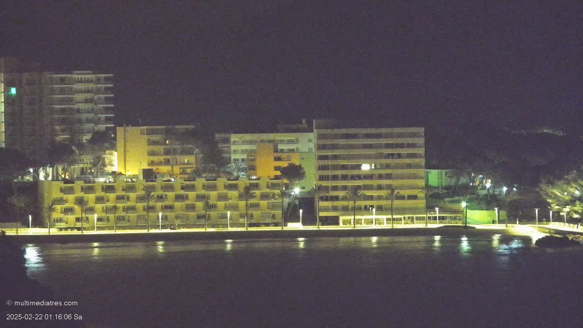 A row of illuminated beachfront buildings at night, reflected in calm water.