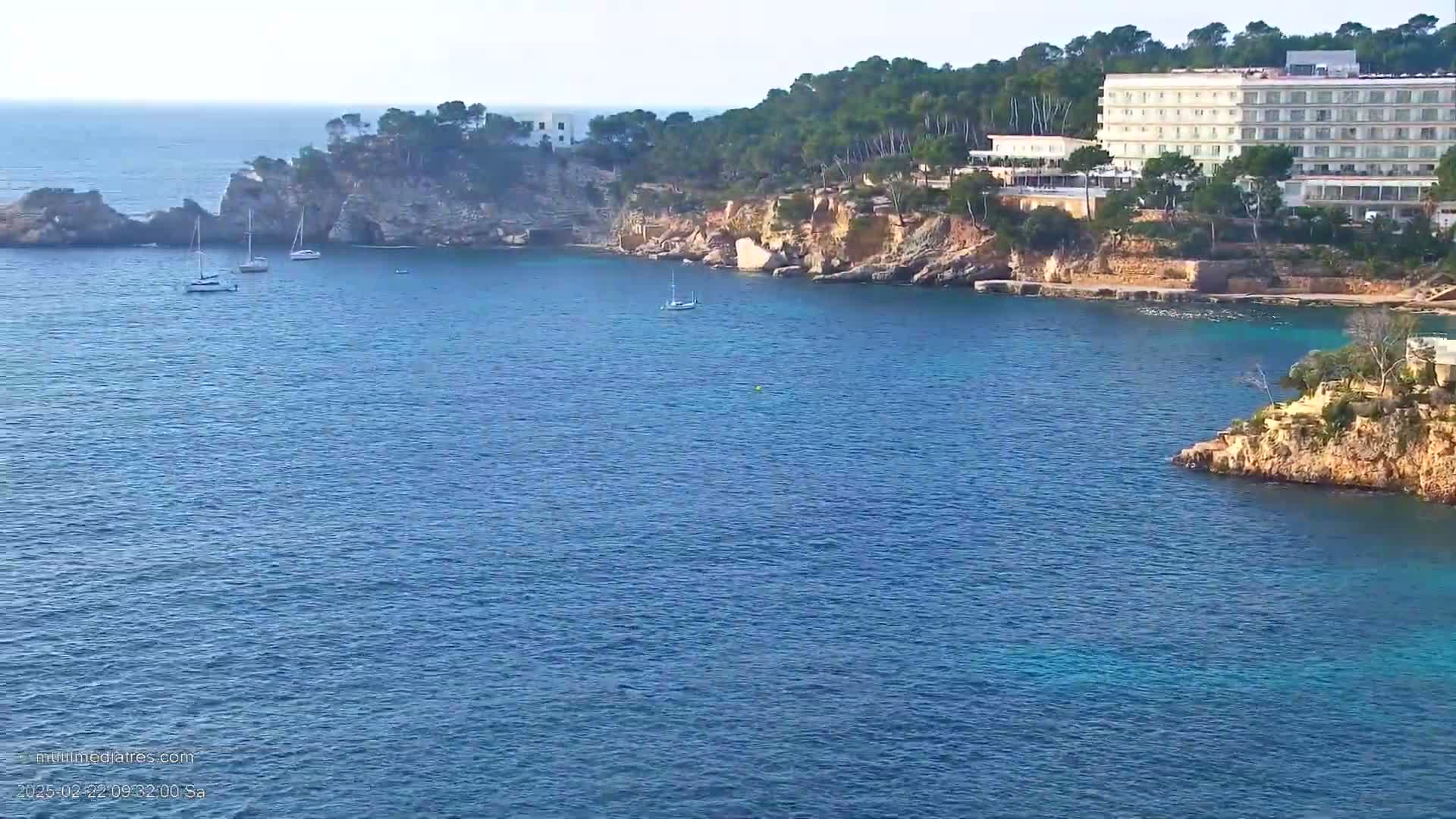 A large hotel sits on a rocky coast overlooking a calm, blue sea with several sailboats.