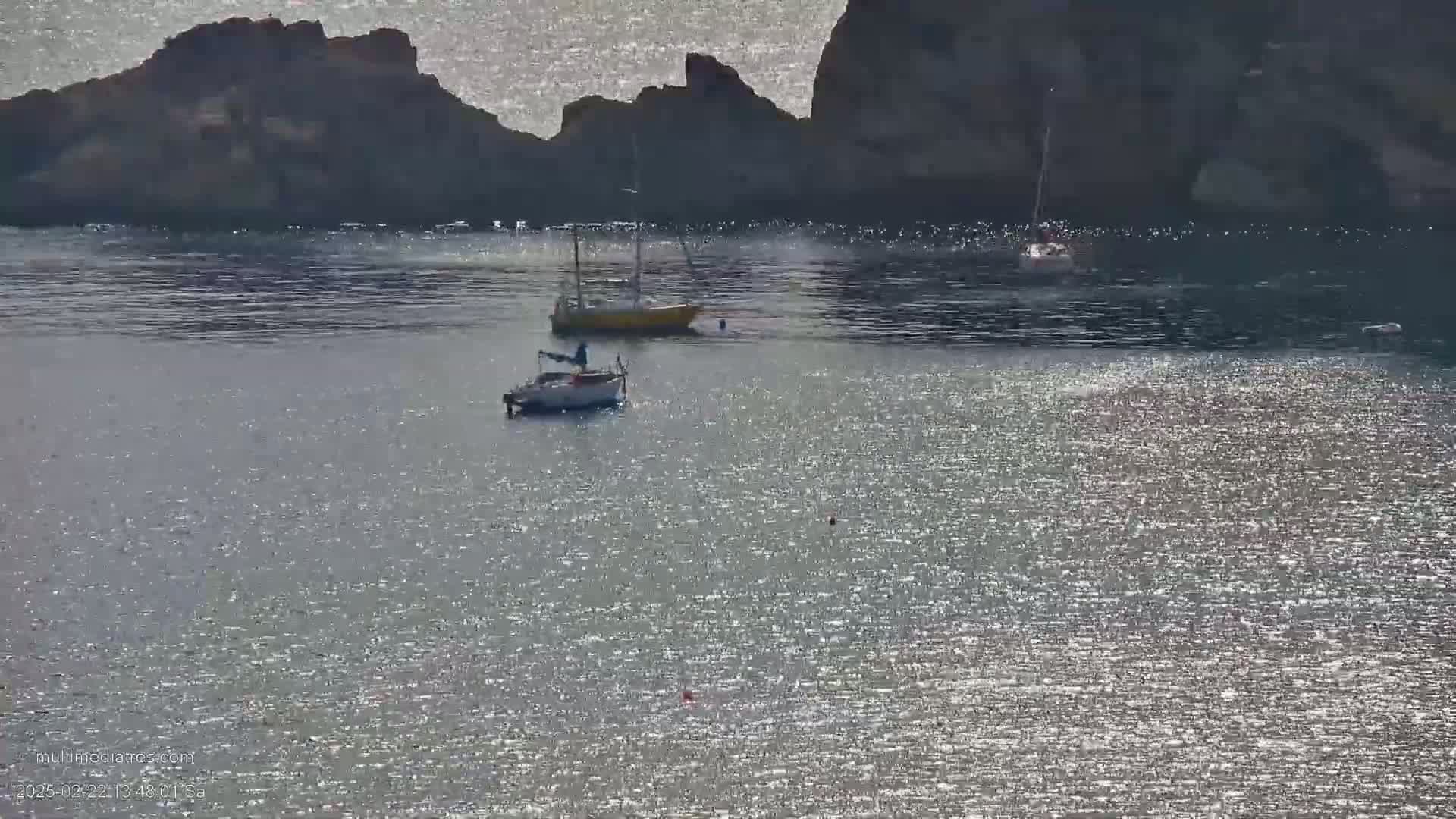 Three small sailboats are moored in a calm, sunlit body of water near a rocky shoreline.