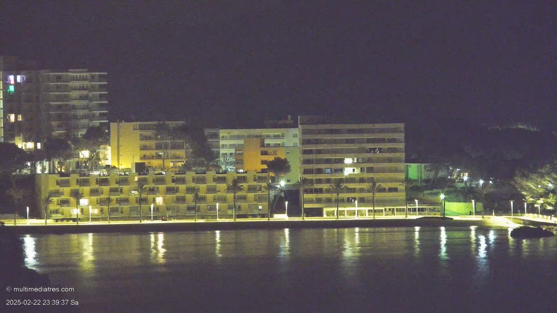 Illuminated apartment buildings line a waterfront at night, their lights reflecting on calm water.